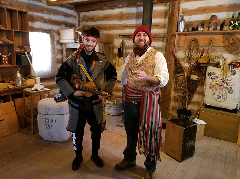 Two men are standing in an old log cabin wearing voyageur costumes