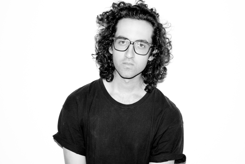 A black and white portrait of Reza Rezai, who has shoulder length, wavy, almost curly dark hair and wears glasses. He is wearing a black t-shirt.