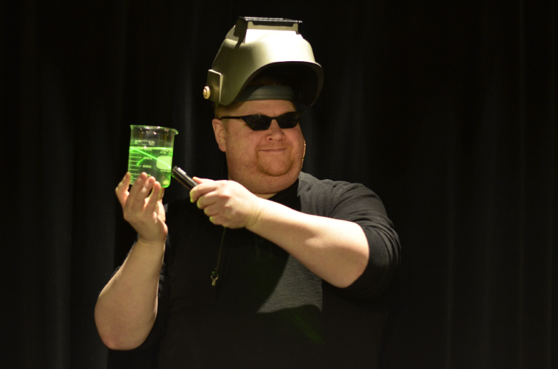 A person with a welding mask points at a beaker filled with a green liquid.
