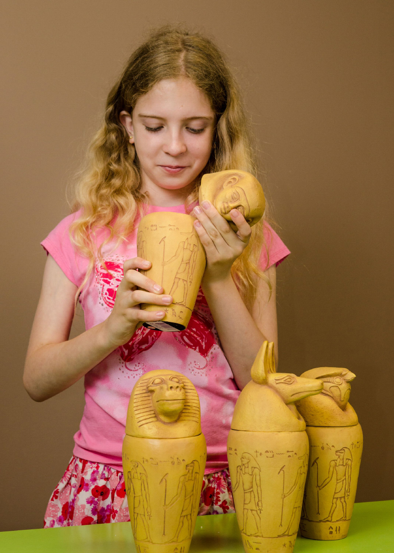 A young child opens an Egyptian urn, in front of her 3 more Egyptian urns are on a green desk.